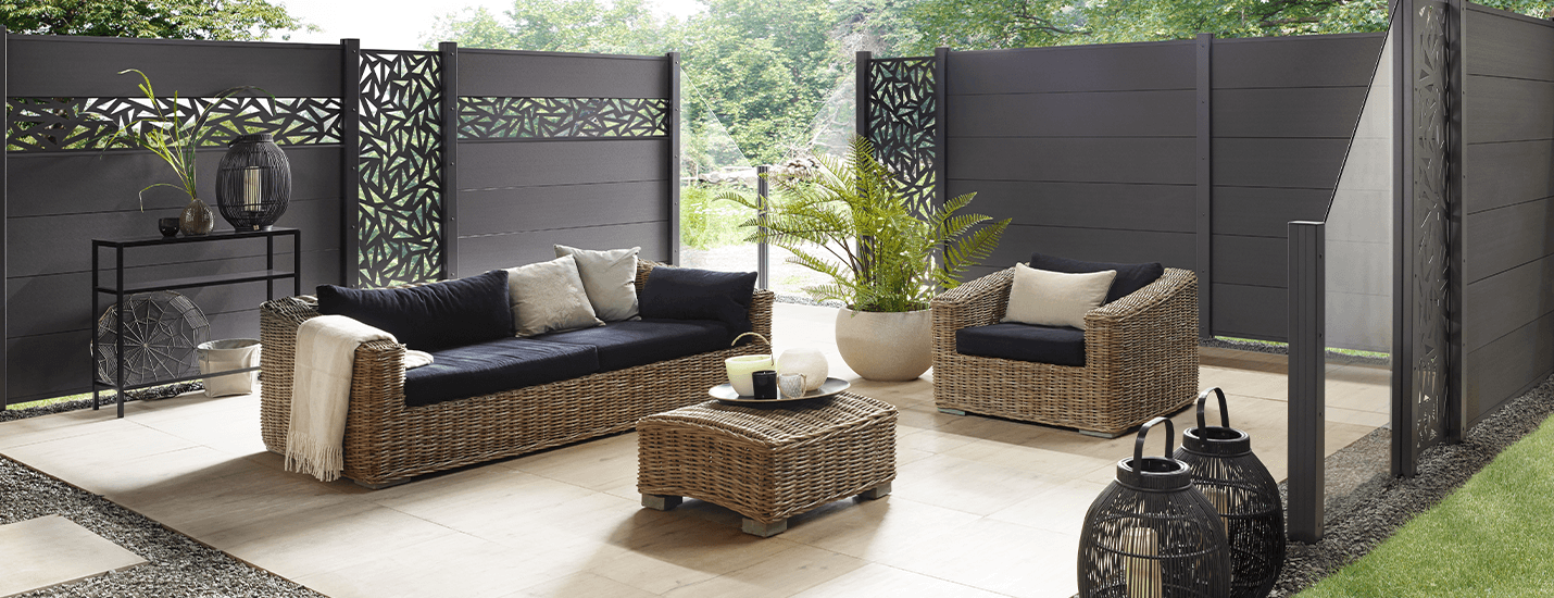 Terrasse mit modernem dunkelbraunem Sichtschutz, unterbrochen mit Glaselementen. Helle Terrassendielen und darauf Korbmöbel und Dekoelemente.